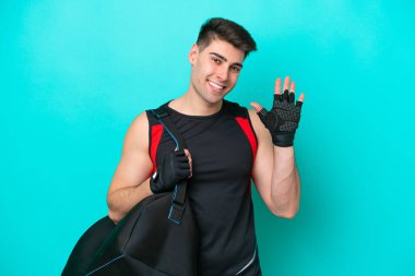 Spor çantalı beyaz bir genç adam mavi arka planda izole edilmiş mutlu bir ifadeyle selam veriyor.