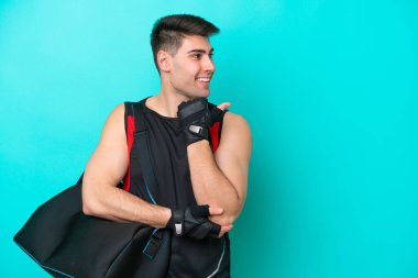 Mavi arka planda izole edilmiş spor çantalı beyaz genç bir adam bir ürünü sunmak için kenarı işaret ediyor.