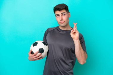 Kafkasyalı genç futbolcu mavi arka planda izole edilmiş ve şans diliyor.