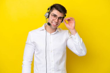 Tele pazarlamacı beyaz adam sarı arka planda gözlüklü ve mutlu bir kulaklıkla çalışıyor.