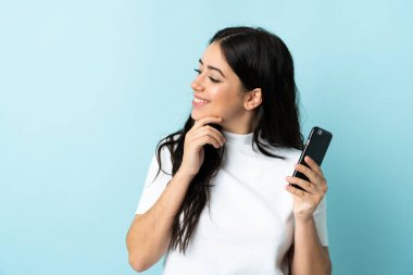 Mavi arka planda izole edilmiş cep telefonu kullanan genç bir kadın bir fikir ve bir bakış açısı düşünüyor.