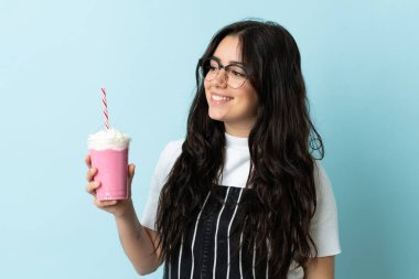 Çilekli milkshake 'i olan genç bir kadın mavi arka planda izole edilmiş, yana bakıyor ve gülümsüyor.
