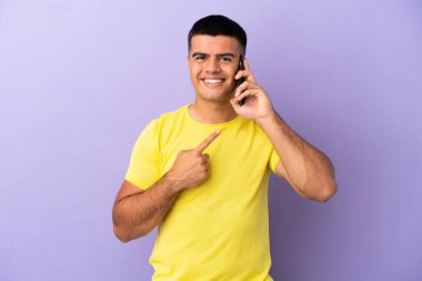 İzole edilmiş mor arka planda cep telefonu kullanan genç yakışıklı bir adam bir ürünü sunmak için yan tarafı işaret ediyor.
