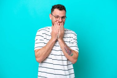 Genç Brezilyalı bir adam mavi arka planda izole edilmiş mutlu ve gülümseyen elleriyle ağzını kapatıyor.