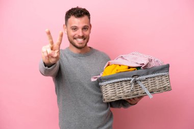 Genç Brezilyalı bir adam pembe arka planda izole edilmiş bir sepet tutuyor gülümsüyor ve zafer işareti gösteriyor.