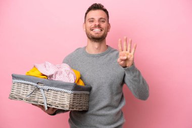 Genç Brezilyalı bir adam pembe arka planda izole edilmiş bir sepet tutuyor mutlu ve parmakları olan dört kişi sayıyor.