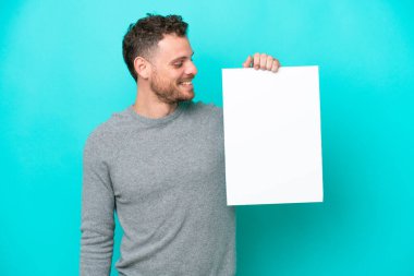 Elinde boş bir levha tutan genç Brezilyalı bir adam mavi arka planda izole edilmiş boş bir levha tutuyor ve ona bakıyor.