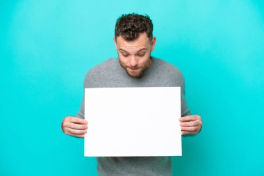Elinde boş bir levha tutan genç Brezilyalı bir adam mavi arka planda izole edilmiş boş bir levha tutuyor ve ona bakıyor.