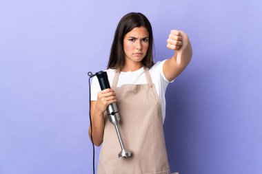 Mor arka planda izole edilmiş el blenderi kullanan Brezilyalı kadın, baş parmağını negatif ifadeyle aşağı gösteriyor