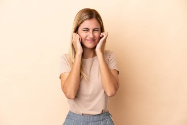 Sarışın Uruguaylı kız bej arka planda izole edilmiş hayal kırıklığına uğramış ve kulaklarını kapatmış.