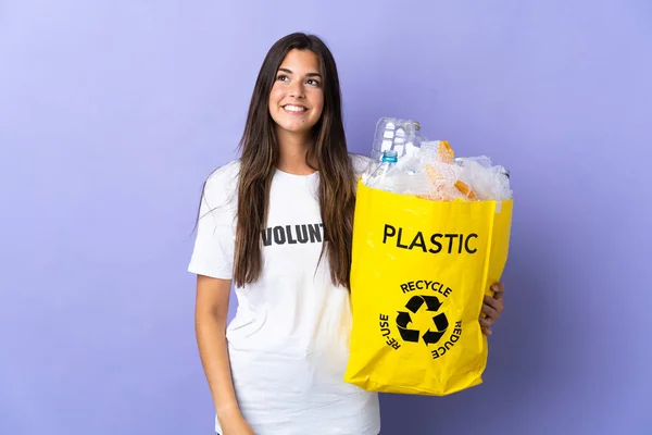紫の背景に隔離されたリサイクルのためのペットボトルの袋を持っている若いブラジル人女性は 上を見上げながらアイデアを考えます — ストック写真