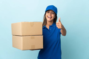 Genç teslimatçı kadın mavi arka planda izole edildi. Başparmakları havada çünkü iyi bir şey oldu.