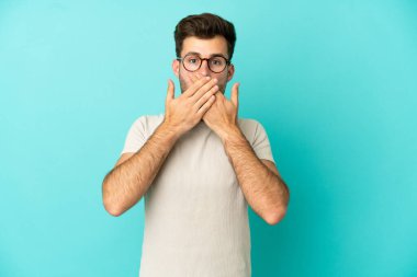 Beyaz tenli yakışıklı adam mavi arka planda izole edilmiş, ağzı elleriyle kapatıyor.