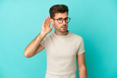 Beyaz tenli yakışıklı adam mavi arka planda izole edilmiş bir şekilde elini kulağına koyarak bir şeyler dinliyor.