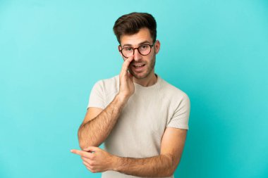 Mavi arka planda soyutlanmış beyaz tenli yakışıklı bir adam bir ürünü sunmak ve bir şeyler fısıldamak için kenarı işaret ediyor.