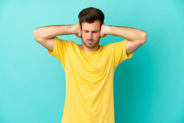 Kafkasyalı yakışıklı adam mavi arka planda izole edilmiş hayal kırıklığına uğramış ve kulaklarını kapatmış.