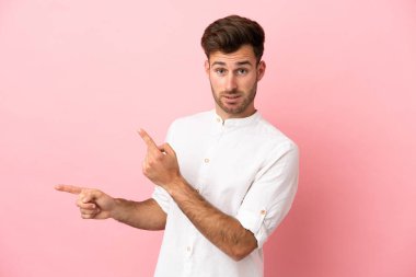 Pembe arka planda izole edilmiş genç, beyaz, yakışıklı bir adam. Korkmuş ve kenarı işaret ediyor.