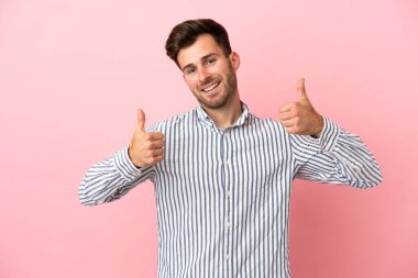 Pembe arka planda izole edilmiş beyaz, yakışıklı bir genç baş parmak hareketi yapıyor.