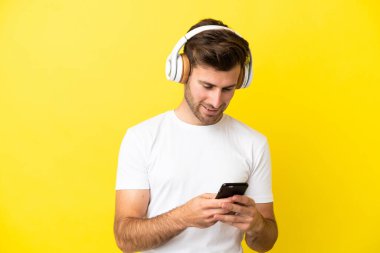 Beyaz tenli yakışıklı adam sarı arka planda izole edilmiş müzik dinliyor ve cep telefonu arıyor.