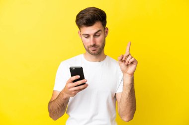Sarı arka planda izole edilmiş genç beyaz adam cep telefonu kullanıyor ve parmağını kaldırıyor.