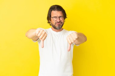 Kıdemli Hollandalı adam sarı arka planda izole edilmiş, baş parmağını iki eliyle aşağı gösteriyor.