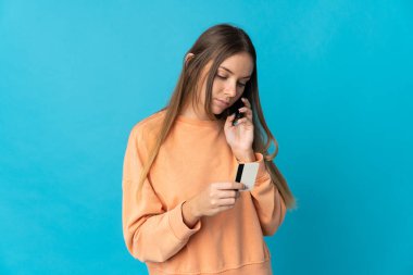 Genç Litvanyalı bir kadın mavi arka planda izole edilmiş ve kredi kartıyla cep telefonuyla alışveriş yapıyor.