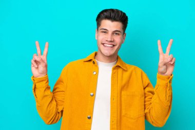 Mavi bakkalda izole edilmiş yakışıklı beyaz adam iki eliyle zafer işareti gösteriyor.