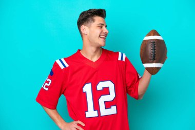 Rugby oynayan beyaz bir adam mavi arka planda izole edilmiş, kalçasında kollarıyla poz verip gülümsüyor.