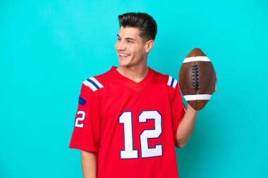 Genç beyaz adam ragbi oynuyor. Mavi arka planda izole edilmiş. Yan tarafa bakıyor ve gülümsüyor.