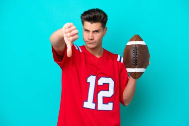 Genç beyaz adam rugby oynuyor. Mavi arka planda izole edilmiş. Negatif bir ifadeyle baş parmağını gösteriyor.