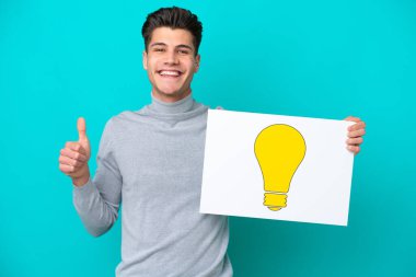 Genç, beyaz bir adam mavi bakkal sahasında izole edilmiş. Elinde başparmağı havada bir ampul simgesi olan bir pankart var.