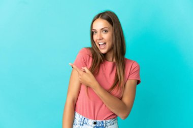 İzole edilmiş mavi arka planı olan genç bir kadın şaşırmış ve işaret ediyor.
