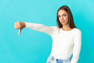 İzole edilmiş mavi arka planda genç bir kadın baş parmağını negatif ifadeyle aşağı gösteriyor.