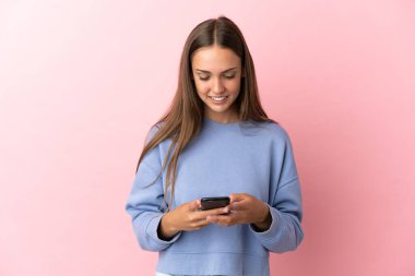 İzole edilmiş pembe arka planı olan genç bir kadın cep telefonuyla mesaj gönderiyor.