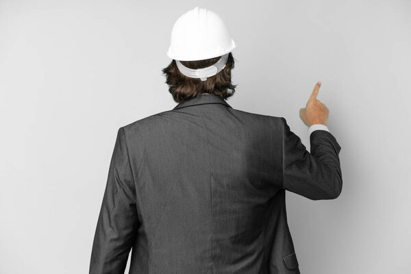 Young architect man with helmet over isolated background pointing back with the index finger