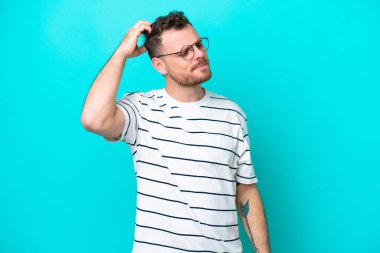 Genç Brezilyalı bir adam mavi arka planda izole edilmiş, kafası kaşınırken şüpheleri var.