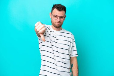 Mavi arka planda izole edilmiş genç Brezilyalı adam, olumsuz ifadeyle baş parmağını aşağı gösteriyor.