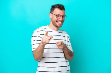 Mavi arka planda izole edilmiş genç Brezilyalı adam şaşırmış ve ön tarafı işaret ediyor.