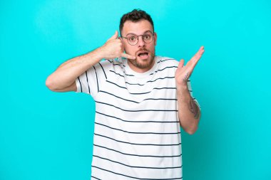 Genç Brezilyalı bir adam mavi arka planda izole edilmiş telefon jestleri yapıyor.