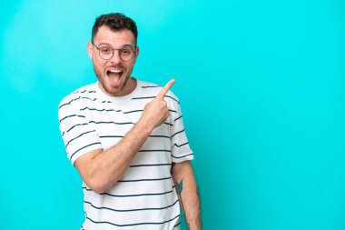 Mavi arka planda izole edilmiş genç Brezilyalı adam şaşırmış ve işaret ediyor.