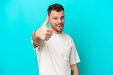 Genç Brezilyalı adam mavi arka planda izole edildi. Başparmakları havada çünkü iyi bir şey oldu.