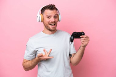 Pembe arka planda izole edilmiş bilgisayar oyunu kontrolörüyle oynayan Brezilyalı genç adam davet etmek için ellerini yana uzatıyor.