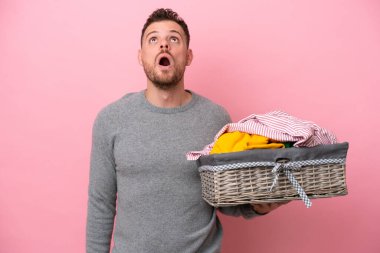 Genç Brezilyalı bir adam pembe arka planda izole edilmiş bir sepet tutuyor yukarı bakıyor ve şaşırmış bir ifadeyle