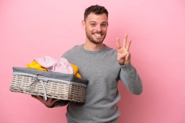 Genç Brezilyalı bir adam pembe arka planda izole edilmiş bir sepet tutuyor mutlu ve parmaklarıyla üçe kadar sayıyor.