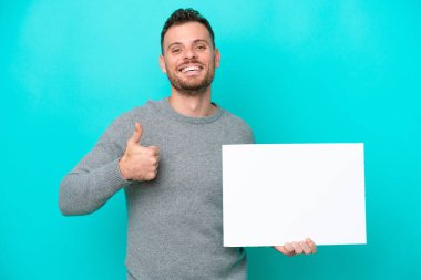Elinde boş bir levha tutan genç Brezilyalı adam mavi arka planda izole edilmiş ve başparmağı havada boş bir pankart tutuyor.