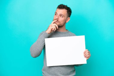 Elinde boş bir levha tutan Brezilyalı genç bir adam mavi arka planda izole edilmiş boş bir levha tutuyor ve düşünüyor: