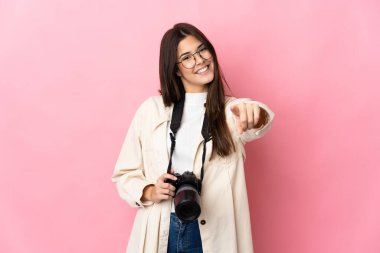 Genç fotoğrafçı Brezilyalı kız pembe arka planda izole edilmiş mutlu bir ifadeyle ön tarafı işaret ediyor.