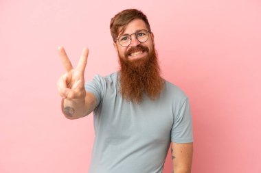 Pembe arka planda izole edilmiş genç beyaz adam gülümsüyor ve zafer işareti gösteriyor.