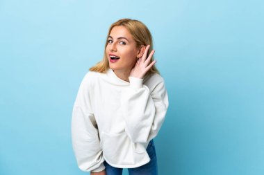 Genç Uruguaylı sarışın kadın mavi arka planda elini kulağına koyarak bir şeyler dinliyor.