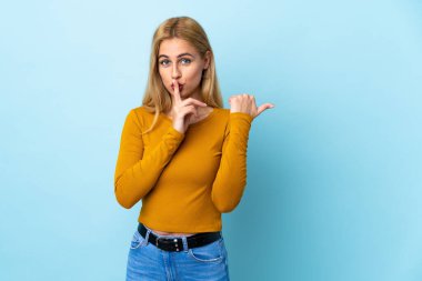 Genç Uruguaylı sarışın kadın, izole edilmiş mavi arka planda. Yan tarafı işaret ediyor ve sessizlik hareketi yapıyor.